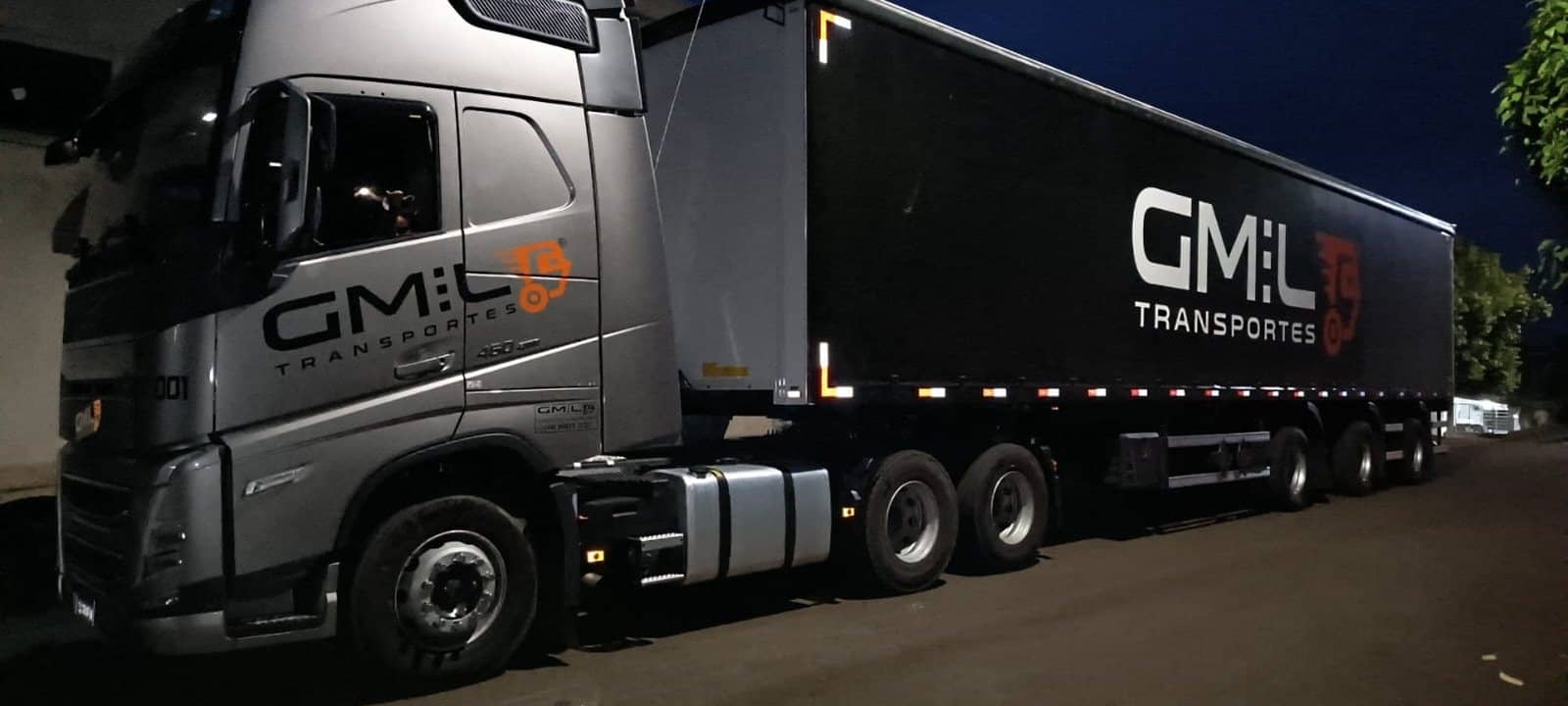 Cavalo Volvo prata da GMIL com carreta sider de lona preta e o logo GMIL, à noite